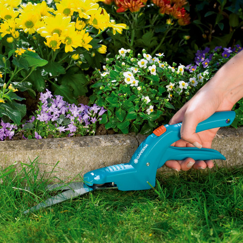 A hand uses blue gardening shears to trim bright-green grass along a colourful flower bed with yellow, purple, and white blooms, conveying a peaceful gardening scene.
