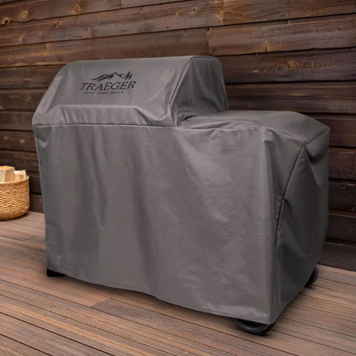 Grey Traeger barbecue grill cover fitted over a Woodridge wood pellet grill on a wooden deck, with Traeger logo printed on the front.