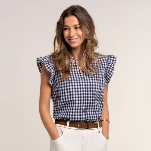 Woman wearing a navy and white gingham sleeveless blouse with frilled shoulders and button front, styled with cream trousers and a brown belt.