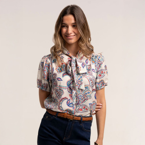 Woman wearing a cream paisley print blouse with short sleeves and tie neck detail, styled with dark indigo jeans and a brown woven belt.