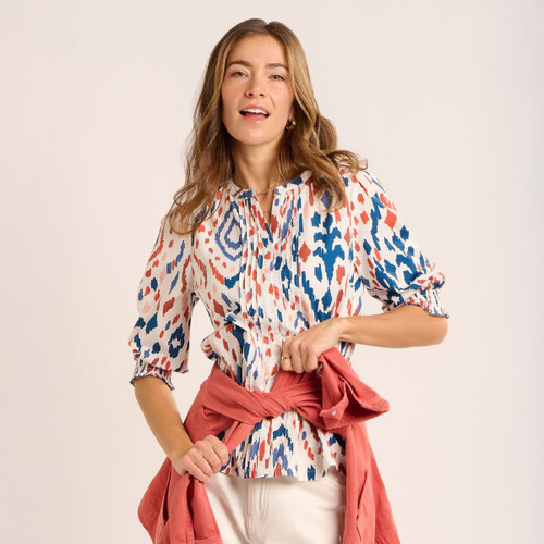 Woman wearing a cream blouse with blue and coral abstract print and three-quarter length sleeves, styled with off-white trousers and a coral jacket tied around the waist.