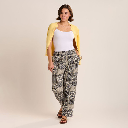 Woman wearing navy and cream patterned wide-leg trousers with a white vest top and yellow cardigan draped over her shoulders, styled with brown sandals against a neutral studio background.