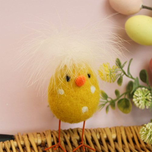 Yellow woollen chick decoration with white polka dots, fluffy feather tuft, orange beak and wire legs, standing against an Easter background.