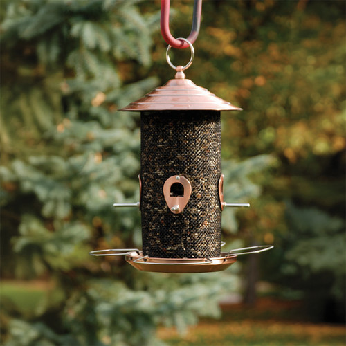 Brushed copper mesh bird feeder filled with mixed seed, hanging outdoors in a garden setting with blurred greenery in the background.