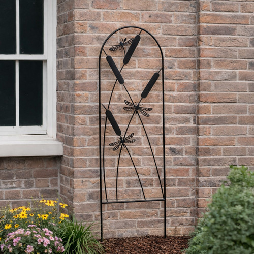 Black metal garden trellis with cattail and dragonfly design standing in a flower bed against a brick wall.