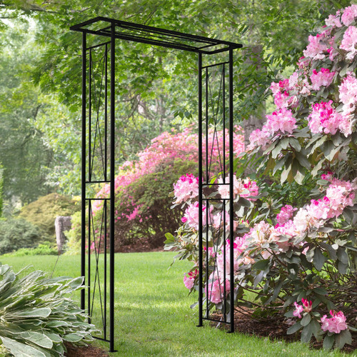 Black metal garden archway standing on a lawn, surrounded by green trees and pink flowering shrubs.