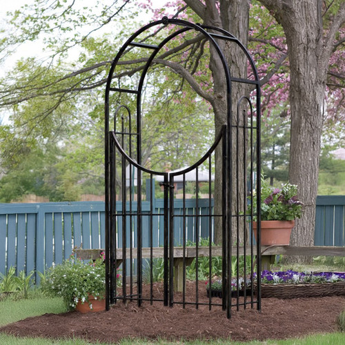 Black metal garden arch with integrated double gates installed in a landscaped garden, surrounded by trees, flowers and a blue fence.