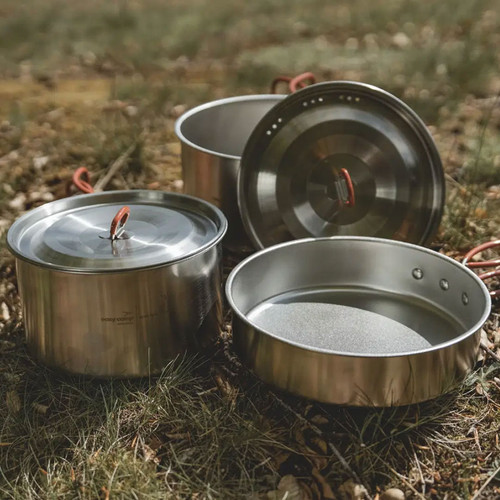 Stainless steel camping cookware set with two pots, lids and frying pan arranged on grass outdoors.