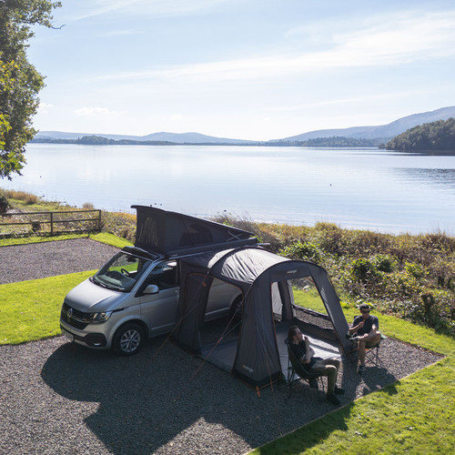 A camper van with an attached tent overlooks a serene lake surrounded by mountains. Two people relax in chairs, enjoying the peaceful, sunny atmosphere.