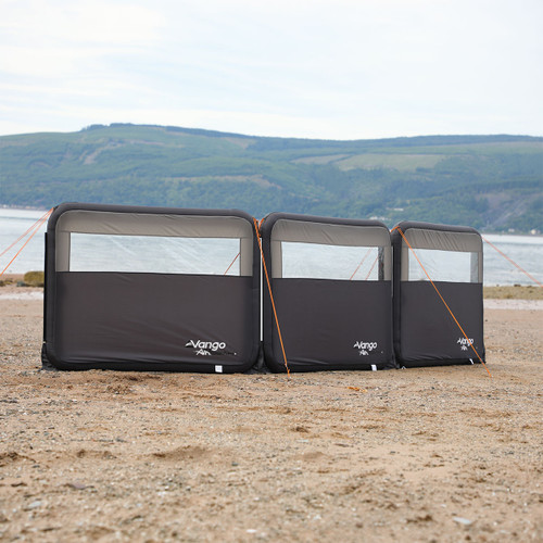Three dark grey inflatable windbreak panels with transparent sections, marked "Vango Air," are set up on a sandy beach, with hills and water in the background. Bright orange guy lines secure them, creating a practical and serene outdoor setting.