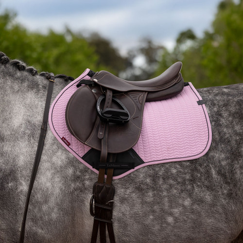 A brown leather saddle on a dappled grey horse with a pink quilted saddle pad. The background features blurred green foliage and a cloudy sky.