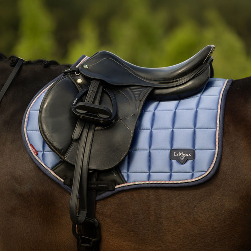 A black saddle rests on a blue, quilted LeMieux saddle pad on a brown horse. The background is a blurred mix of green hues, evoking calmness.