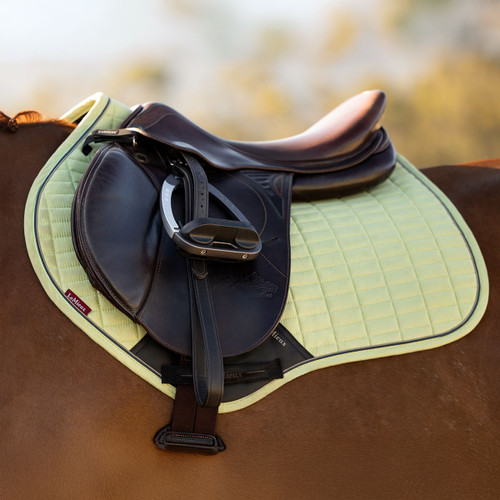 A close-up of a dark brown leather horse saddle on a light green quilted saddle pad, placed on a brown horse with a blurred outdoor background.