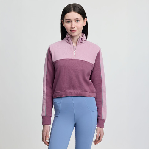 A woman wearing a mauve and pink quarter-zip pullover and blue leggings stands against a plain background. She has long dark hair and a subtle smile.