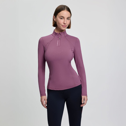 A woman in a fitted mauve long-sleeve top with a high collar and zipper stands against a plain grey background, conveying a sleek, athletic style.