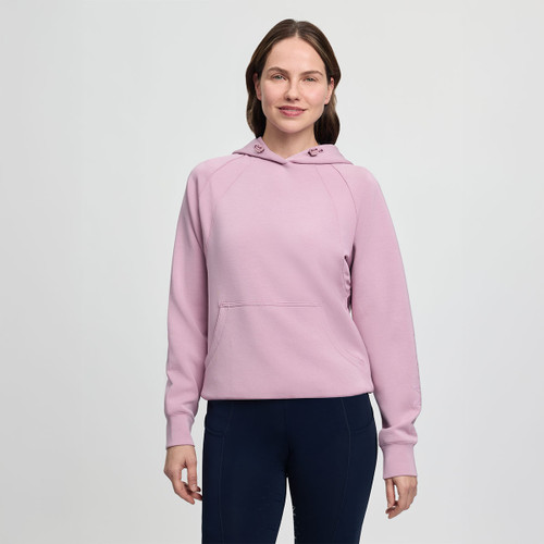 A woman stands smiling in a light pink hoodie and dark pants against a plain gray background. Her relaxed pose conveys a casual and comfortable mood.