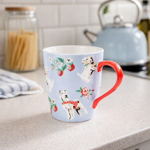 Light blue ceramic mug with a bright red handle, decorated with playful dog illustrations, strawberries and small flowers. The mug sits on a kitchen countertop with a pasta jar, tea towel and blue kettle softly blurred in the background.
