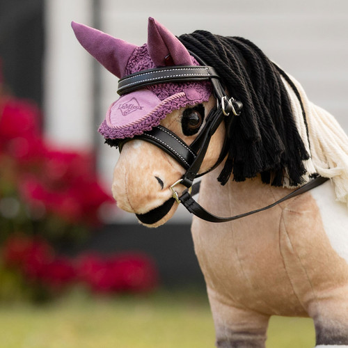 A plush toy horse wearing a lavender ear bonnet and black bridle stands in focus. Red flowers and greenery blur softly in the background, conveying a playful tone.