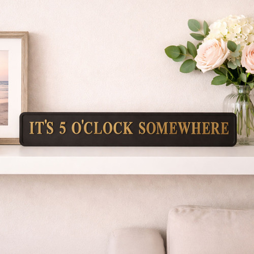Black rectangular sign with gold lettering reading ‘IT’S 5 O’CLOCK SOMEWHERE’ displayed on a white shelf, next to a framed picture and a vase of flowers.
