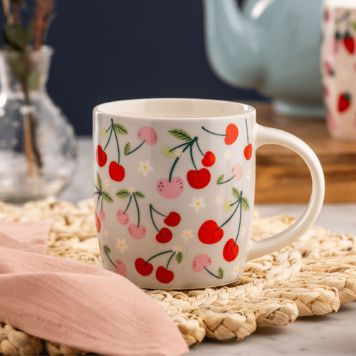 White ceramic mug with red and pink cherry pattern on woven placemat, cozy kitchen scene with pastel teapot and soft fabric