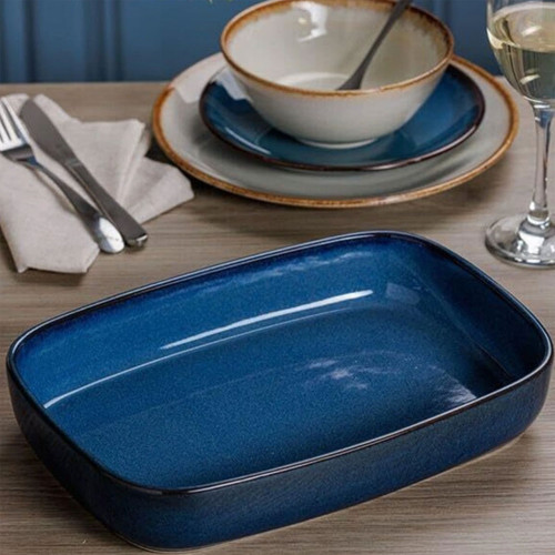 Rectangular blue ceramic baking dish on wooden dining table with plates, bowl, cutlery, and wine glass, modern kitchen table setting