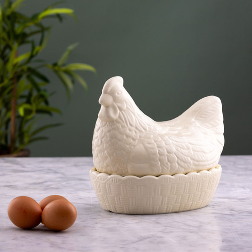 White ceramic hen-shaped egg storage container with lid on marble countertop, two brown eggs beside it, farmhouse kitchen decor