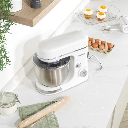 White Salter stand mixer with stainless steel bowl on a kitchen worktop, surrounded by baking ingredients including eggs, flour and a rolling pin.