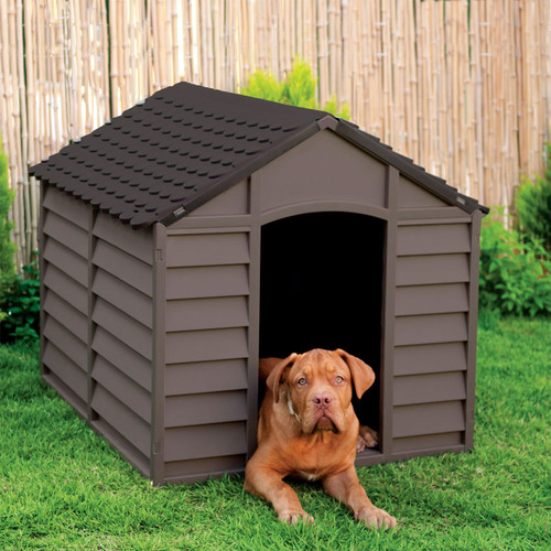 Brown plastic dog kennel with pitched roof, featuring a large dog resting in the doorway on a grassy garden lawn
