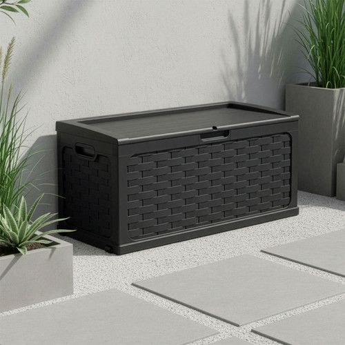 Dark grey rattan-effect outdoor storage box with lid, placed on a modern patio beside planters