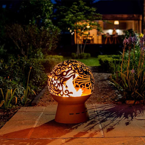 Decorative steel fire globe with intricate dragon cut-out design glowing at night, creating warm ambient light in a landscaped garden.