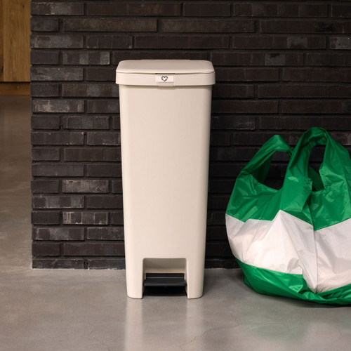 Soft beige Brabantia StepUp pedal bin positioned beside a kitchen wall with reusable shopping bags on the floor.