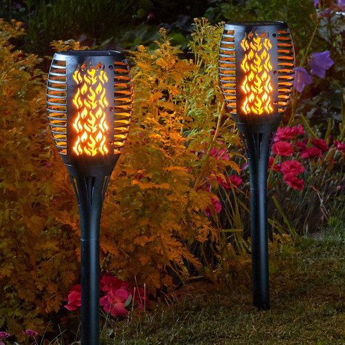 Two black solar garden stake lights with a decorative flame-cut design glow with a warm amber light, creating the appearance of flickering flames. The lights are positioned among colourful flowers and foliage in a garden at dusk.