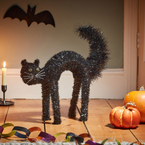 Black tinsel Halloween cat decoration with an arched back and raised tail, displayed on a wooden floor alongside pumpkins and seasonal décor.