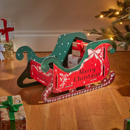 Wooden Christmas sleigh decoration with hand-painted red and green festive design, featuring “Merry Christmas” lettering and used as a decorative gift holder beside a Christmas tree.