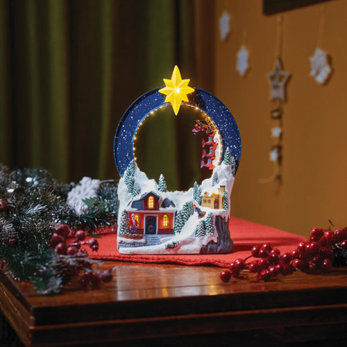 Illuminated Christmas village decoration featuring a snowy winter scene, glowing star topper, lit cottages, and Santa’s sleigh with reindeer suspended within a circular frame.