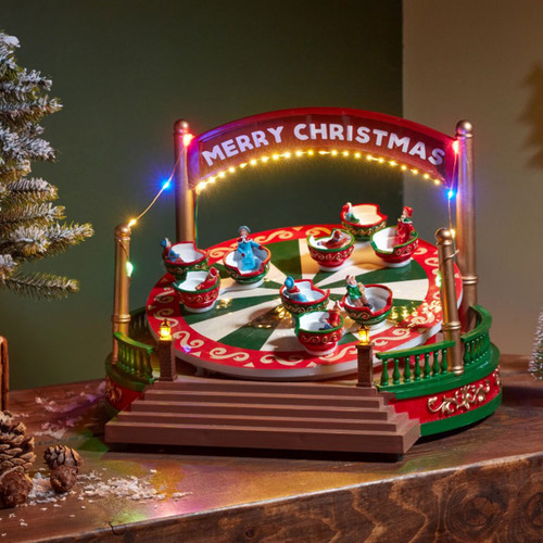 Animated Christmas fairground ride decoration with colourful LED lights, spinning teacup-style seats, festive figures, and a "Merry Christmas" sign, displayed on a tabletop.