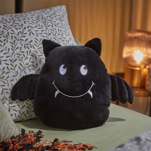 Black plush bat-shaped Halloween cushion with embroidered smile, white fangs, soft wings and ears, displayed on a sofa for seasonal home décor.