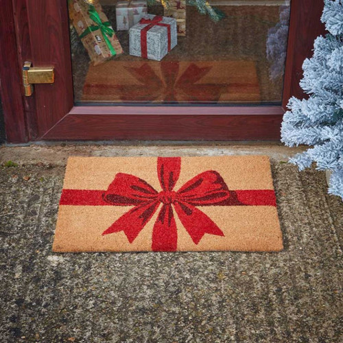 Christmas gift-themed coir doormat with a red ribbon and bow design, placed outside a front door with festive decorations visible through the glass.