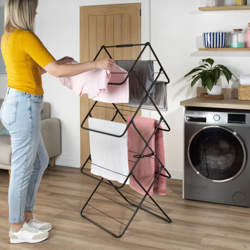 Black multi-tier folding clothes airer in use with towels and clothing drying in a modern laundry room beside a washing machine.