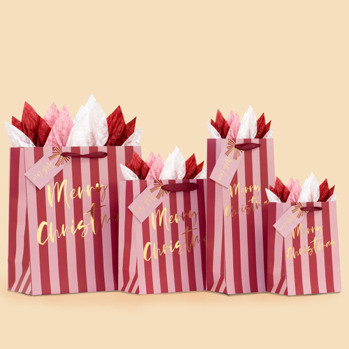 Four Christmas gift bags in assorted sizes, featuring alternating pink and burgundy vertical stripes with gold foil “Merry Christmas” lettering. Each bag has burgundy ribbon handles, a matching gift tag, and red, pink, and white tissue paper, arranged against a warm beige background.
