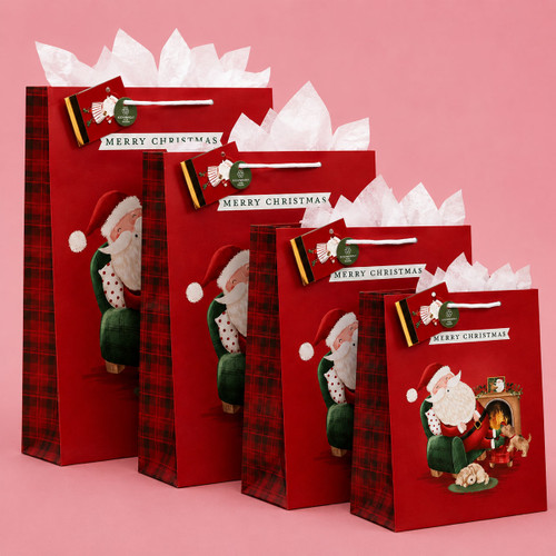 Four red Santa Christmas gift bags in assorted sizes with white tissue paper on a pink background.