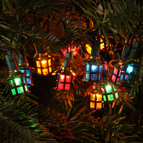 Multicoloured lantern-shaped Christmas string lights glowing among evergreen branches, featuring decorative red, blue, green and amber lanterns for festive indoor or outdoor décor.