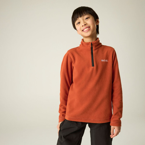 Child wearing a burnt orange half-zip fleece with black hiking trousers, smiling at the camera.