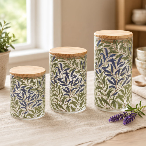Set of three William Morris Willow Bough glass storage canisters in small, medium and large sizes with natural bamboo lids, featuring the iconic blue and green leafy pattern, displayed together on a linen tablecloth.
