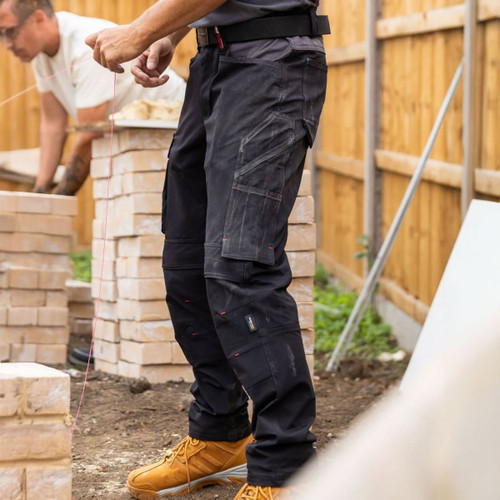 Black TuffStuff work trousers worn on a construction site, featuring reinforced knees, multiple utility pockets and contrasting grey waistband panels.