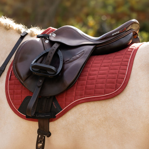 Close-up of a brown English saddle fitted over a red LeMieux quilted saddle pad with contrasting piping on a light-coloured horse.