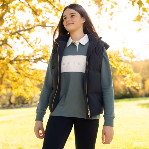 Girl wearing a navy padded LeMieux gilet over a sage green long-sleeve polo top outdoors among golden autumn trees.