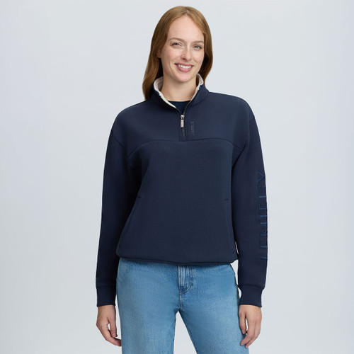 Woman wearing a navy LeMieux quarter-zip sweatshirt with high fleece-lined collar, embroidered sleeve branding and blue jeans.