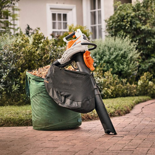 STIHL cordless leaf vacuum with collection bag positioned beside a garden waste bag filled with autumn leaves.