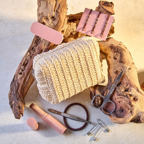 An arranged collection of a woven cream zip bag, pink nail file, rose gold hair pins, scissors, a lip balm, and hair ties on wood.